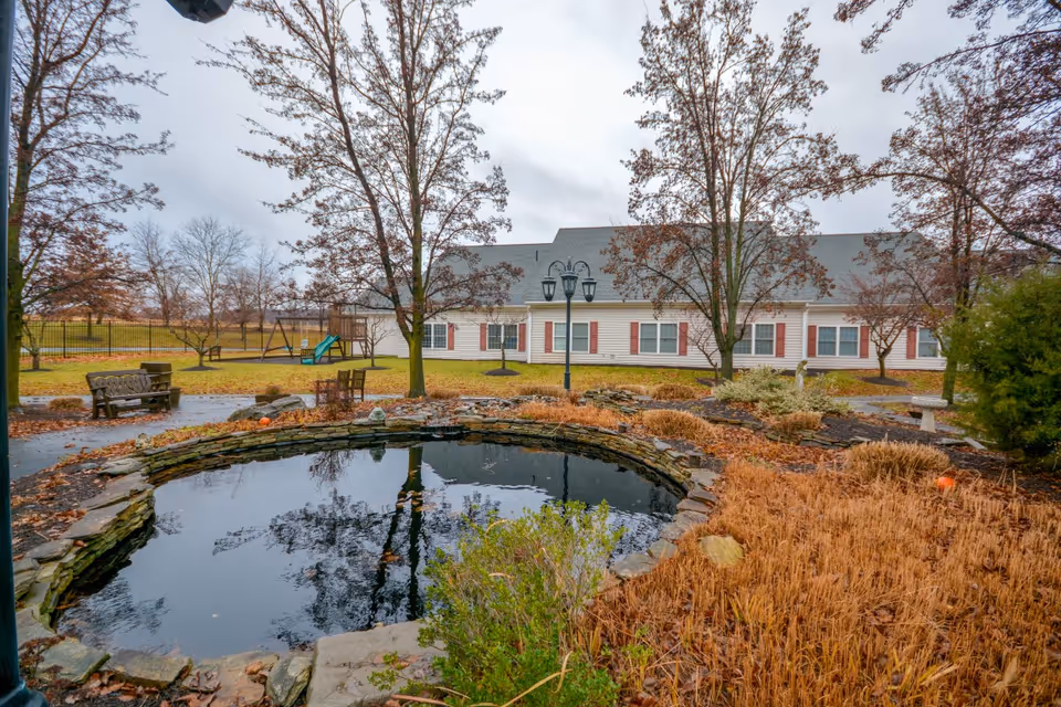 Stone-edged pond and landscaped courtyard with benches and trees in front of a single-story retirement living building.