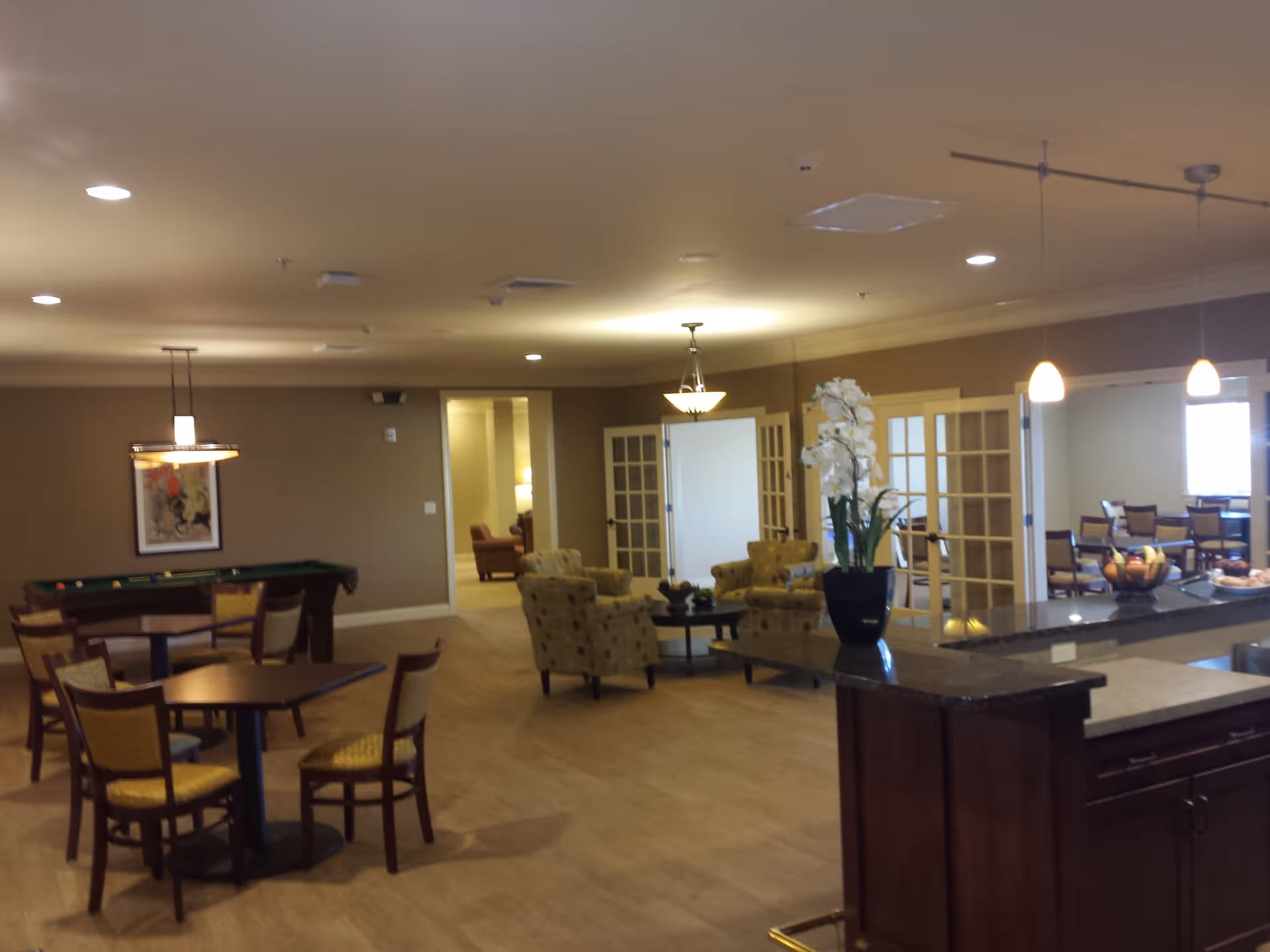 Interior view of a senior living facility common area with tables and chairs, a pool table, comfortable armchairs around a coffee table, and a kitchen counter with a vase of white flowers and fruit bowls. The room has warm lighting and beige walls with French doors leading to another room with more seating.