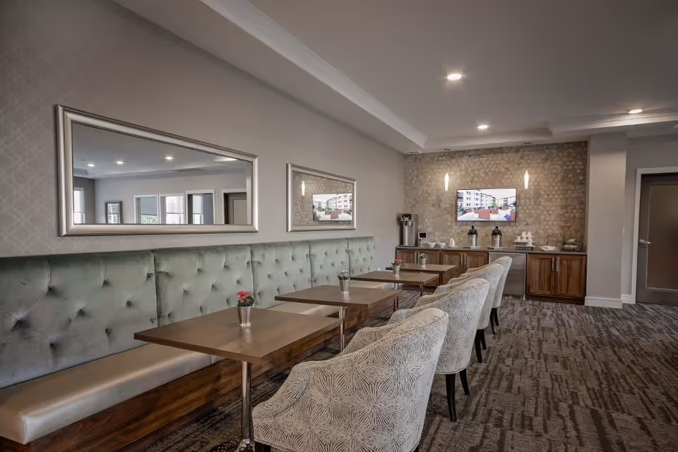Dining/lounge area with tufted banquette seating, tables and upholstered chairs facing a refreshment counter and wall-mounted TV.