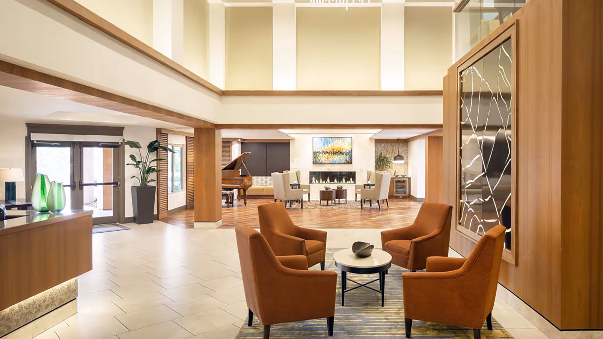 Bright modern lobby with four orange armchairs around a small round table, a reception desk, grand piano, and a seating area by a fireplace.