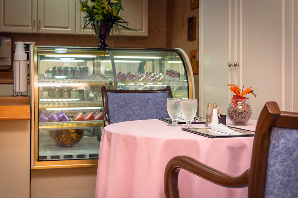 A small dining area with a round table covered with a pink tablecloth, set with two glasses of water, menus, salt and pepper shakers, and a decorative vase with orange flowers. Behind the table is a refrigerated display case containing various beverages and desserts. The setting appears cozy and inviting.