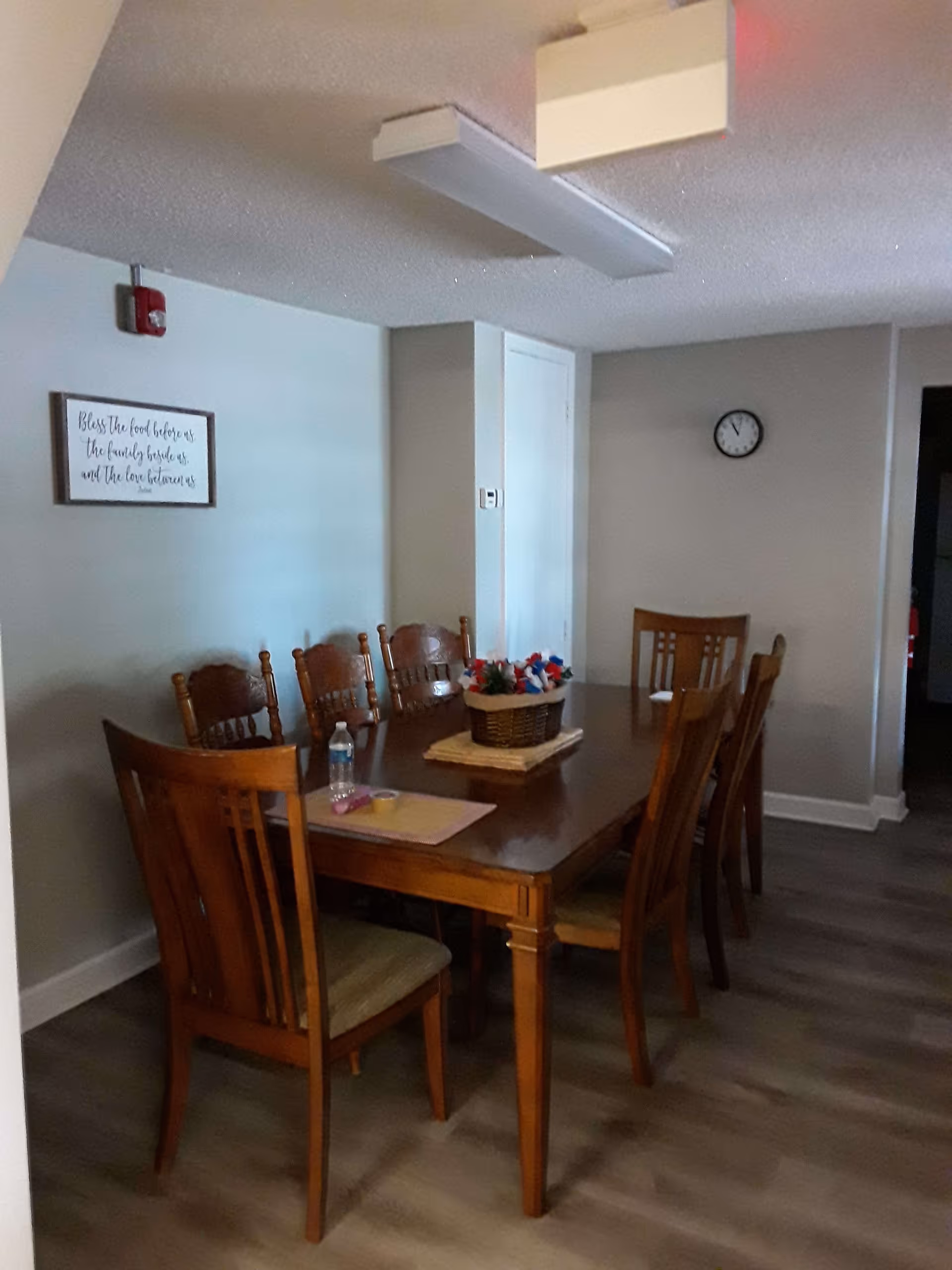 A dining room with a wooden table surrounded by eight wooden chairs. On the table, there is a basket with red, white, and blue flowers, a water bottle, and some small items. The walls are painted light gray, with a framed sign that reads 'Bless the food before us, the family beside us, and the love between us.' A clock is mounted on the far wall, and the floor has wood-style flooring.