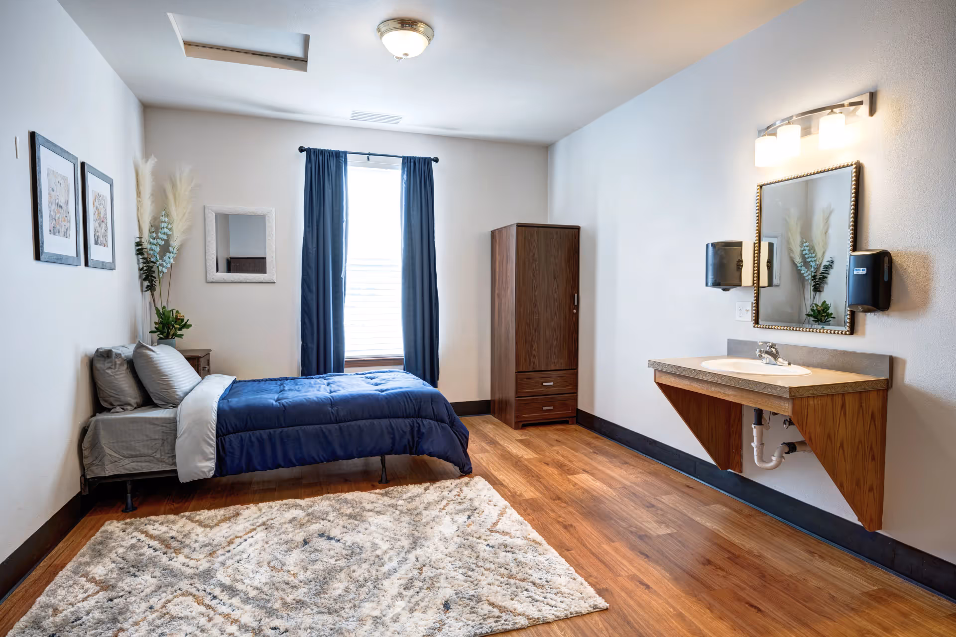 A simple bedroom with a single bed dressed in blue bedding, a wooden wardrobe, a wall-mounted sink and mirror, and a window with dark curtains.