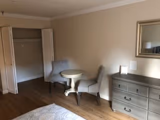 A bedroom with light-colored walls and wooden flooring. There is an open closet with white bi-fold doors on the left side. In the center, there is a small round table with two gray upholstered chairs. On the right side, there is a gray dresser with three drawers, a small lamp, and a rectangular mirror hanging above it.