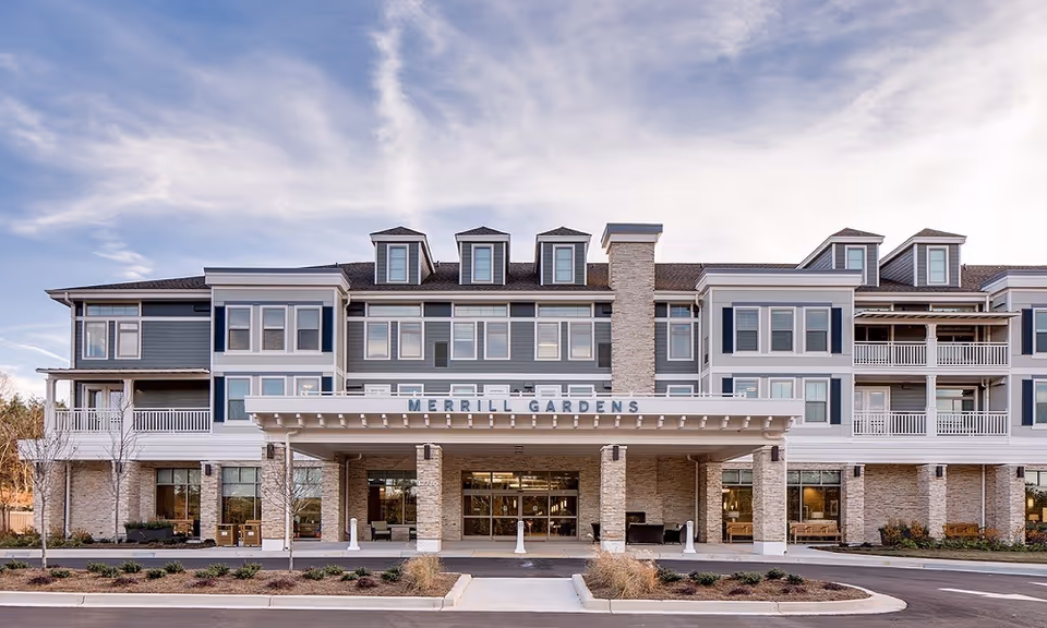 Front exterior view of Merrill Gardens at Woodstock, a three-story senior living facility with a covered entrance, multiple windows, balconies, and a stone and siding facade under a partly cloudy sky.