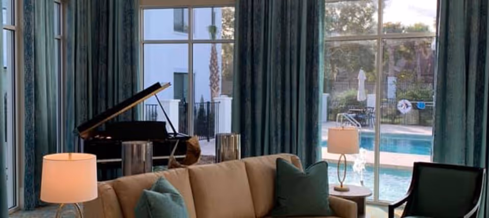 Sunlit living room with a sofa, armchairs, lamps and a grand piano, overlooking a pool through large windows.