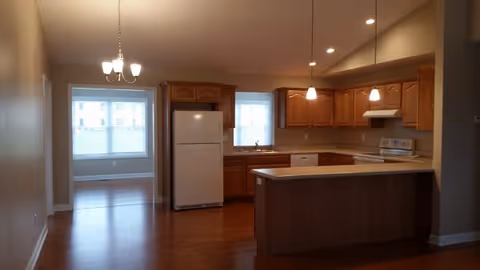 Interior view of a kitchen with wooden cabinets, a white refrigerator, stove, and dishwasher. There is a kitchen island with a countertop and three pendant lights hanging above it. Adjacent to the kitchen is an empty dining area with a chandelier and two windows letting in natural light.