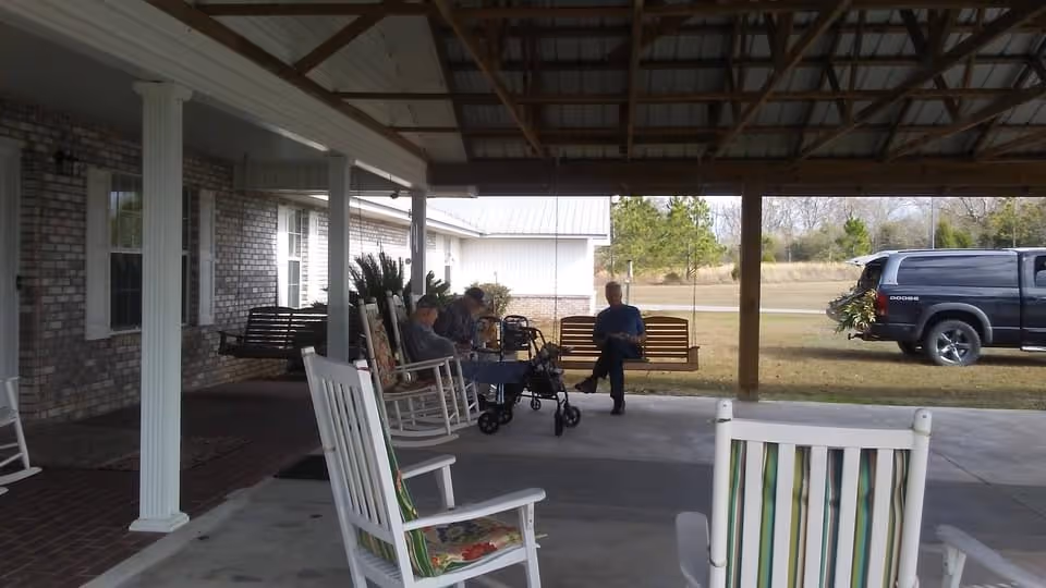 Covered outdoor patio area with rocking chairs and wooden swings. Three elderly people are seated, two on rocking chairs and one on a swing, with a walker nearby. A black Dodge truck is parked on the grass in the background, and trees and open land are visible beyond the patio.