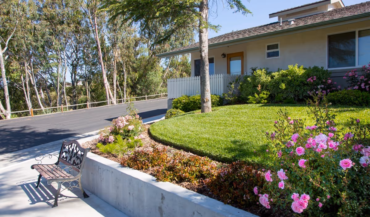 Outdoor garden area at Atterdag Village of Solvang featuring a green lawn, blooming pink flowers, shrubs, a tree, a paved driveway, and a decorative metal and wood bench on a concrete sidewalk.