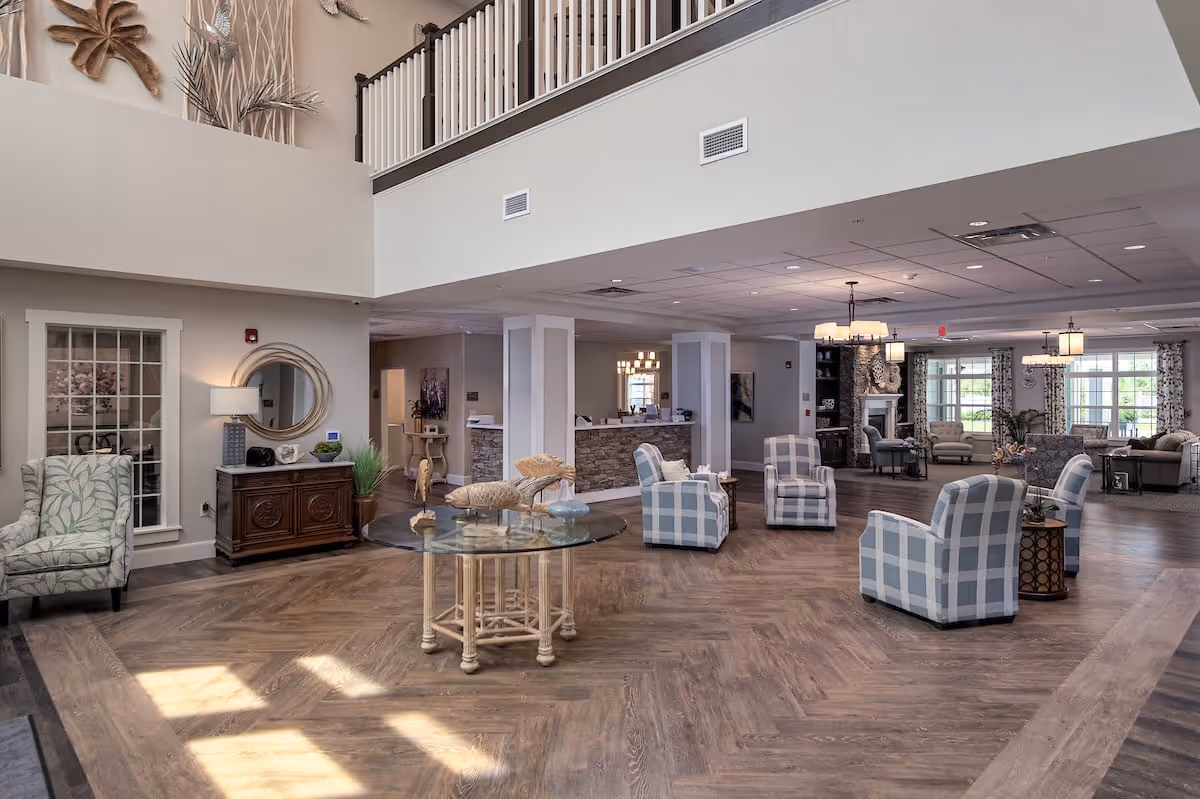 Spacious and well-lit senior living facility common area with wooden flooring, multiple comfortable armchairs in blue and white plaid, a round glass table with decorative items, a wooden sideboard with a lamp and mirror, and large windows allowing natural light. The area includes a reception desk and a lounge area with sofas and a fireplace in the background.