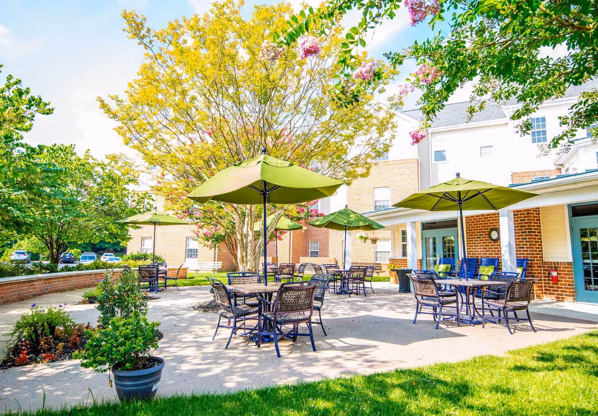 Sunny outdoor patio with tables, chairs, and green umbrellas in front of a brick senior living building.
