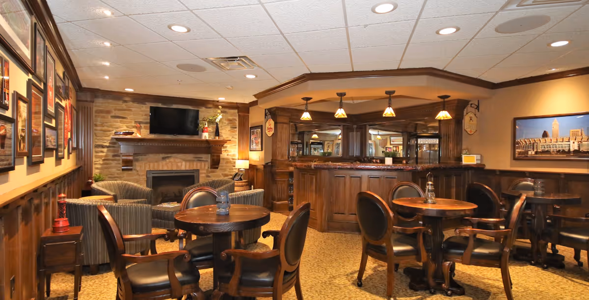 A cozy common area with wooden tables and chairs, a stone fireplace with a mounted TV above it, and a wooden bar with pendant lights hanging from the ceiling. The walls are decorated with framed pictures and a large photo of a city skyline.