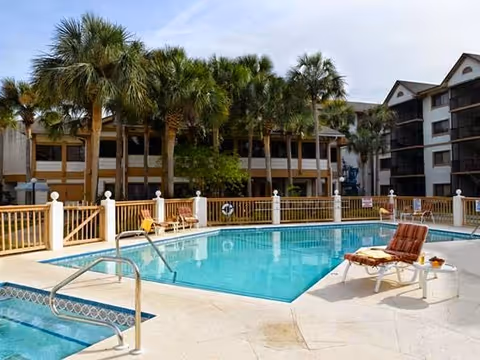 Outdoor swimming pool area at Vitality Living Baypoint Village with lounge chairs and palm trees surrounding the pool. Multi-story residential buildings are visible in the background under a partly cloudy sky.