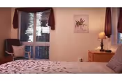 A cozy bedroom with a bed in the foreground, a nightstand and lamp, a cushioned chair by a curtained window, and a framed floral print on the wall.