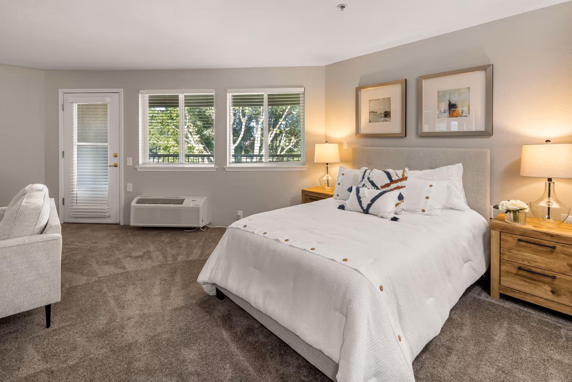 A cozy bedroom in a senior living community featuring a neatly made bed with white bedding and decorative pillows. The room has beige walls, two framed abstract artworks above the bed, two wooden nightstands with lamps, a small vase with flowers, a beige armchair, carpeted floor, and three windows letting in natural light. A door with blinds leads outside.
