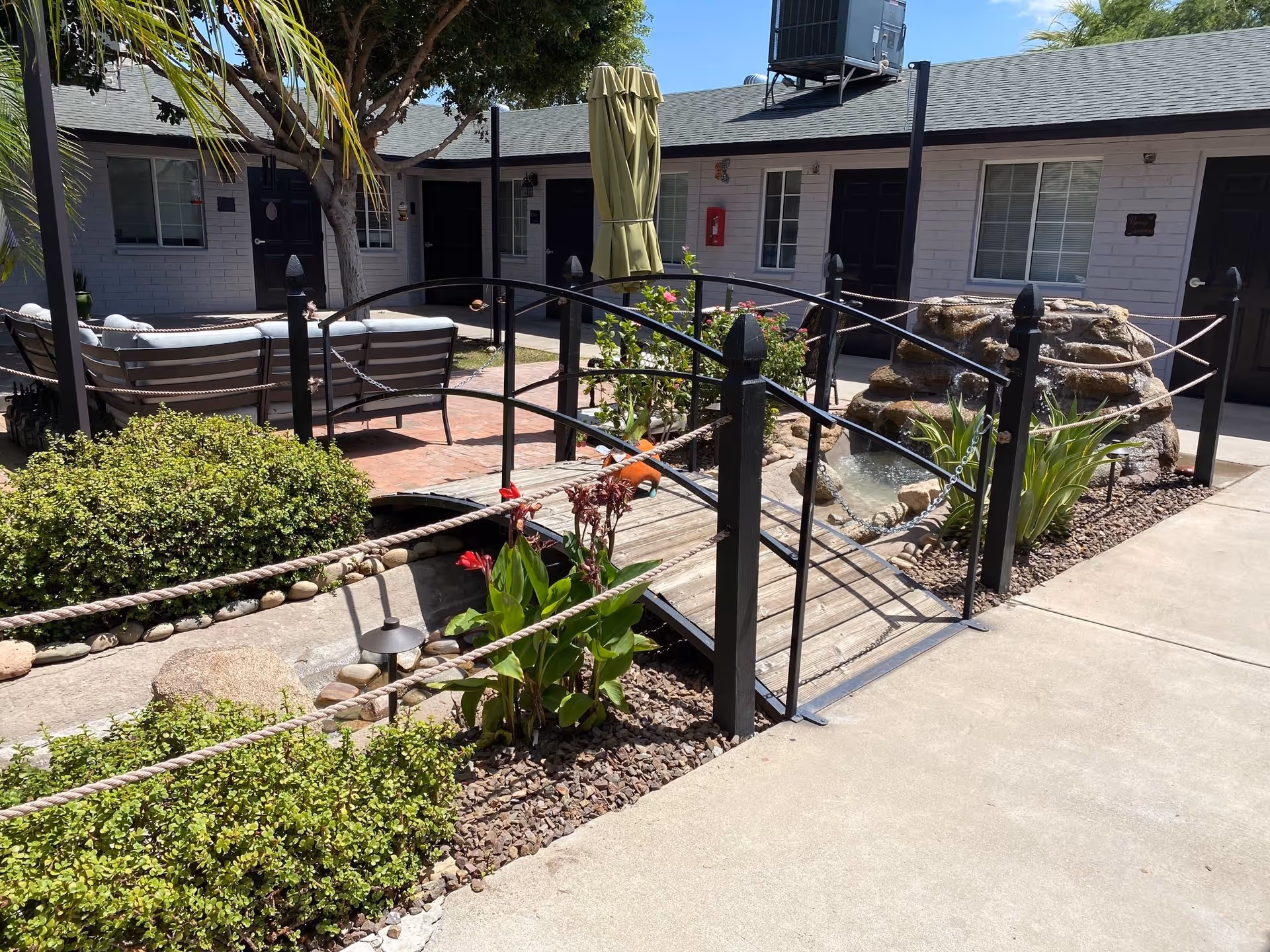 Outdoor courtyard area at Arizona Desert Falls featuring a small wooden footbridge with black metal railings over a landscaped water feature with plants and rocks. There is patio seating with cushioned chairs and a closed umbrella, surrounded by bushes and trees, with building entrances visible in the background under a clear blue sky.