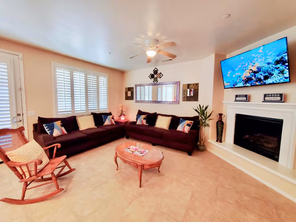A cozy living room with a large dark brown sectional sofa adorned with colorful pillows, a wooden rocking chair with a cushion, a small wooden coffee table with magazines, a wall-mounted flat-screen TV displaying an underwater scene, a white fireplace with decorative items on the mantel, a ceiling fan with a light, and a large mirror on the wall reflecting the room's light.