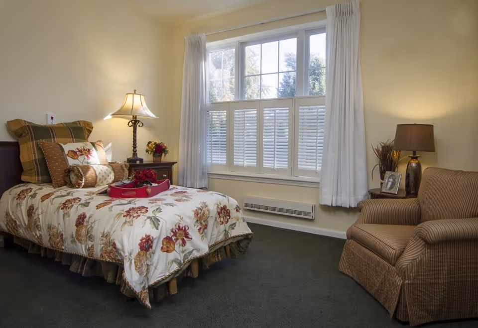 A cozy bedroom with a single bed covered in floral bedding and multiple pillows. There is a wooden nightstand with a decorative lamp and a small flower arrangement. A large window with white curtains and shutters lets in natural light. An upholstered armchair with a striped pattern sits next to another small table with a lamp and framed photo.