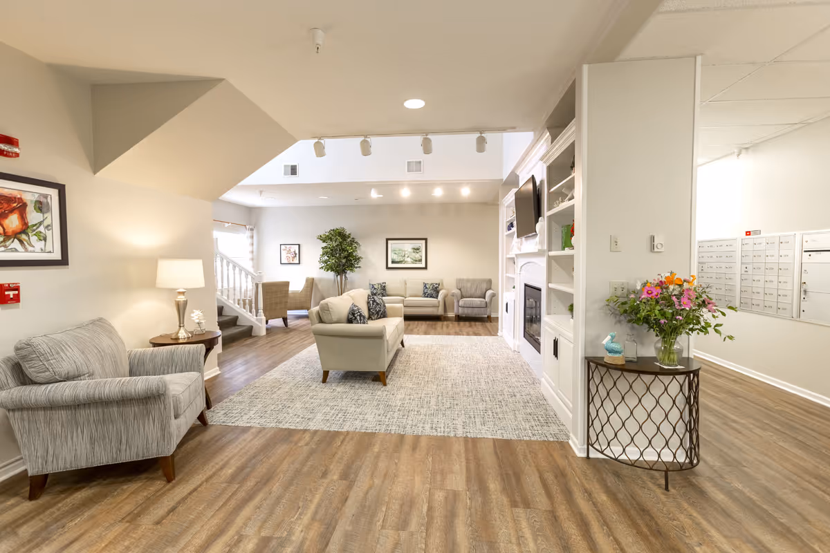 Bright, spacious common lounge with sofas, armchairs, a fireplace and a table with flowers in a senior living facility.