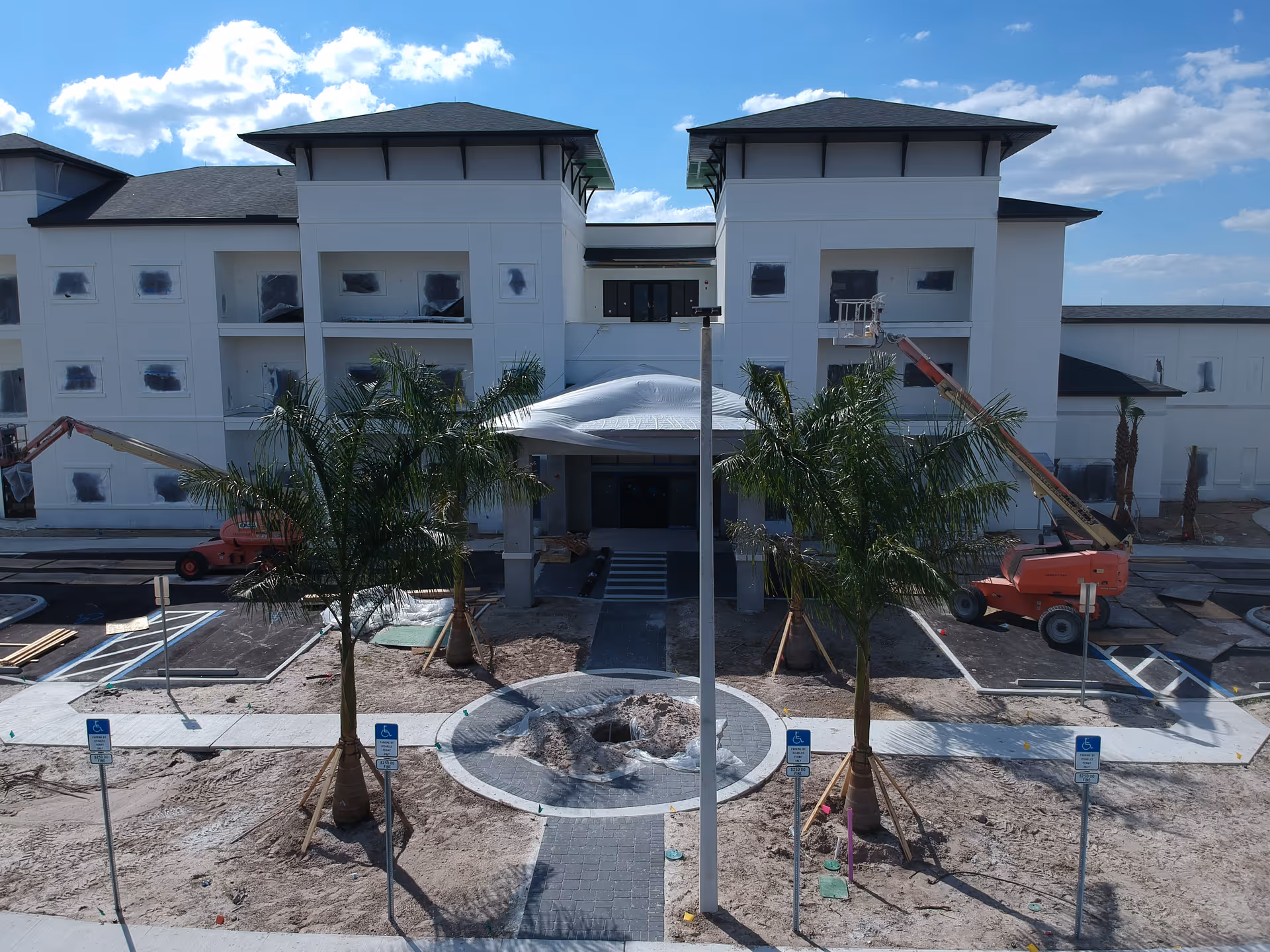 Front exterior view of a large white building under construction with two palm trees and a circular paved area in front. Construction equipment and materials are visible around the site, and the sky is partly cloudy.