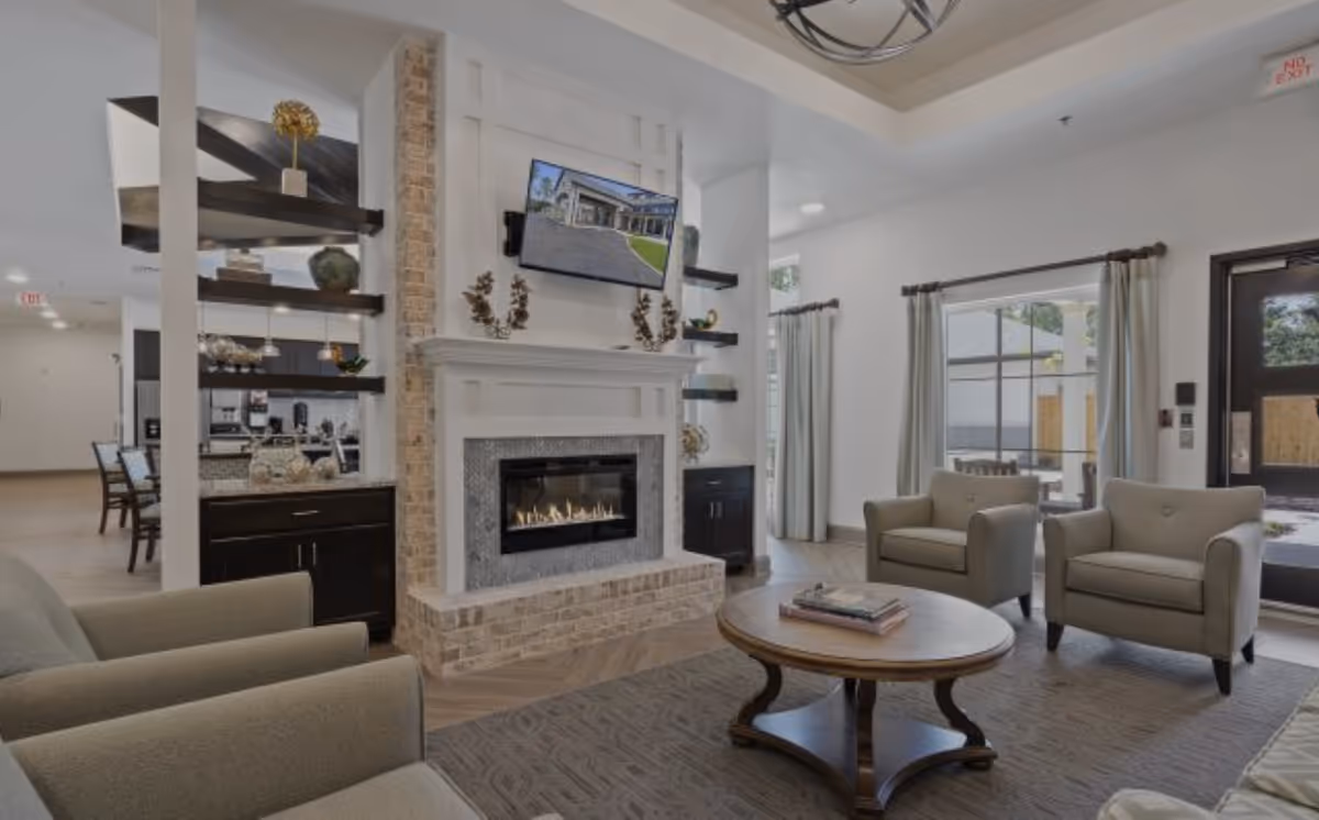 A cozy living room area in a senior living facility featuring a modern fireplace with a TV mounted above it, two beige armchairs, a round wooden coffee table with books, large windows with curtains, and an open view into a dining area and kitchen.