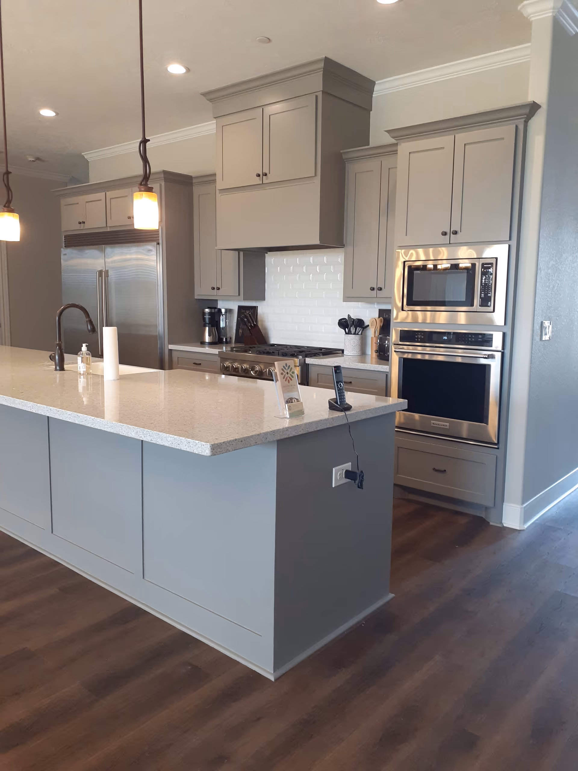 Modern kitchen with gray cabinetry, a large island with a quartz countertop, stainless steel refrigerator, built-in microwave and oven, pendant lighting, and hardwood flooring.