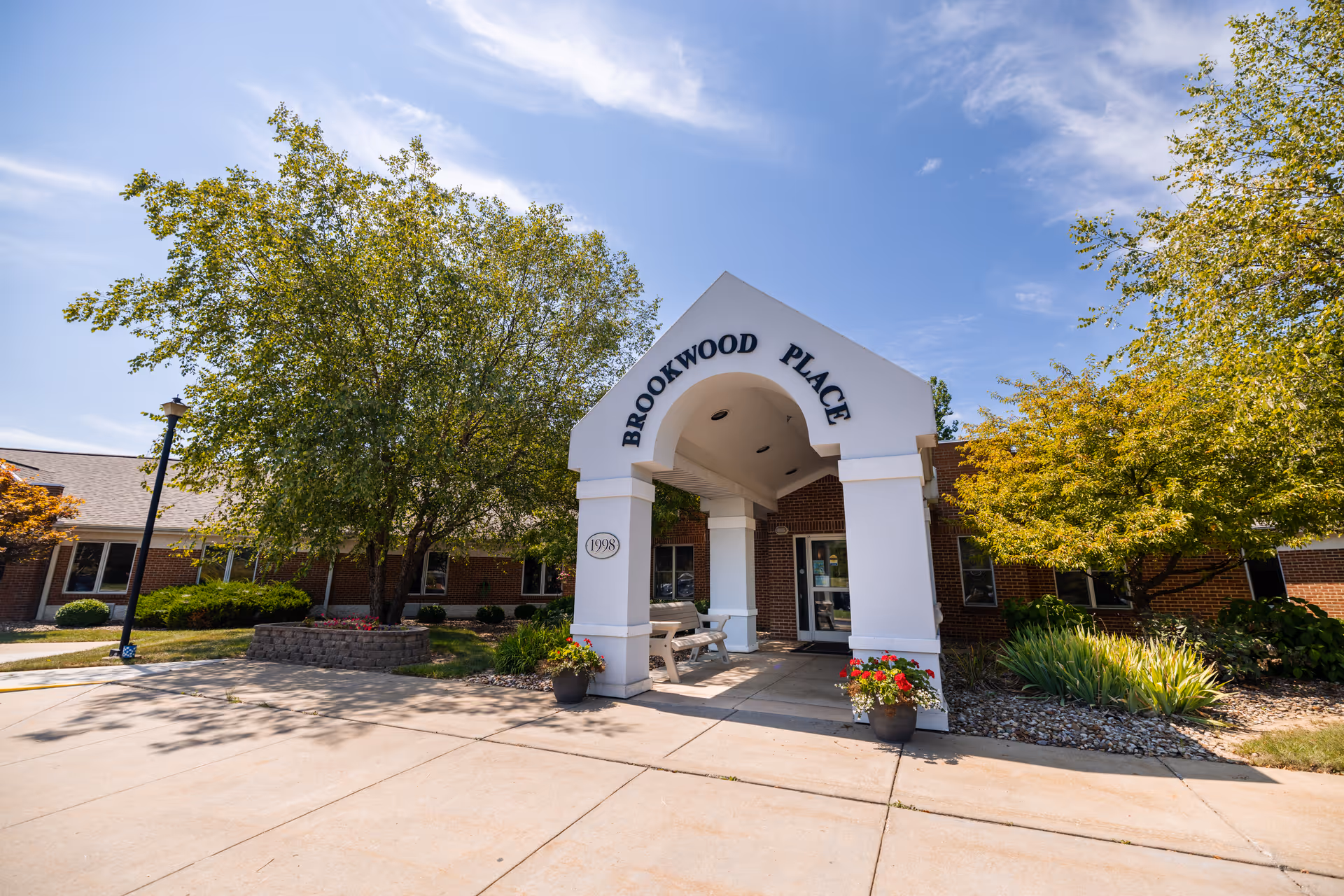 Entrance to Brookwood Place, a single-story brick building with a white archway over the entrance. There are trees and plants around the building and a clear blue sky above.