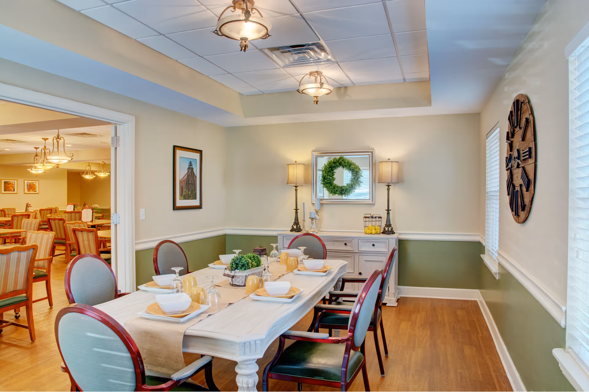 A well-lit dining room with a rectangular table set for six people. The table has a beige runner, white bowls, yellow mugs, and glassware. The room features green and beige walls, wooden flooring, two table lamps on a white sideboard, a decorative wreath on a mirror, and a large wooden wall clock. In the background, another dining area with multiple tables and chairs is visible.