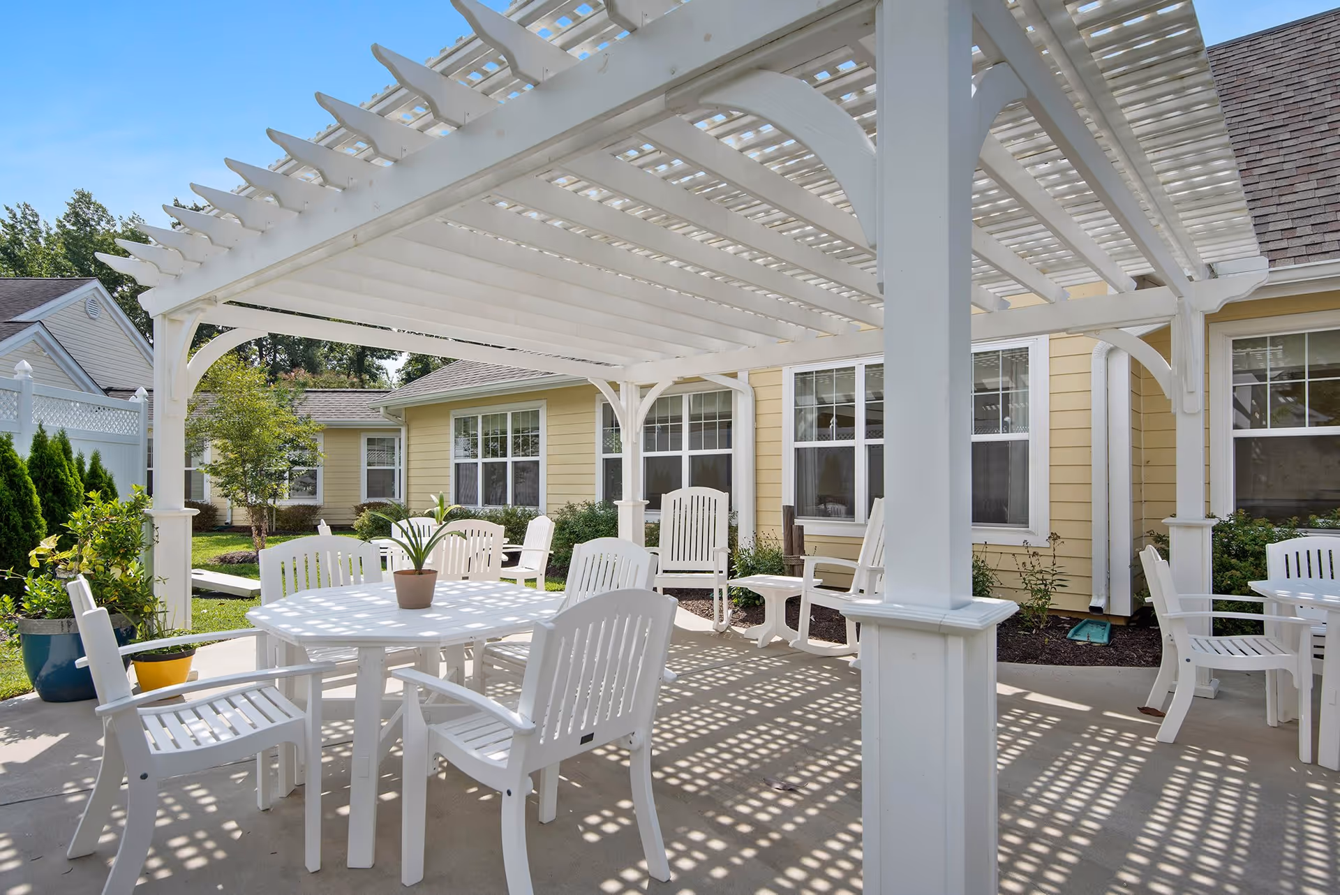Outdoor patio area at Candle Light Cove featuring a white pergola providing partial shade over white tables and chairs. The patio is surrounded by a yellow building with multiple windows and some greenery including potted plants and small trees.
