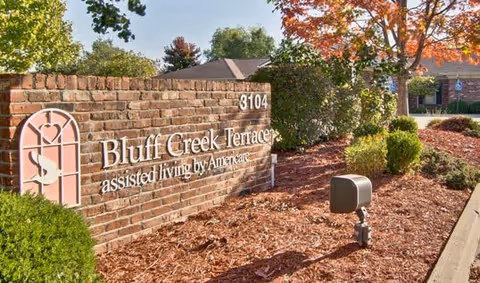 Brick entrance sign reading 'Bluff Creek Terraces assisted living' with landscaped mulch beds, shrubs, and the facility building visible behind it.