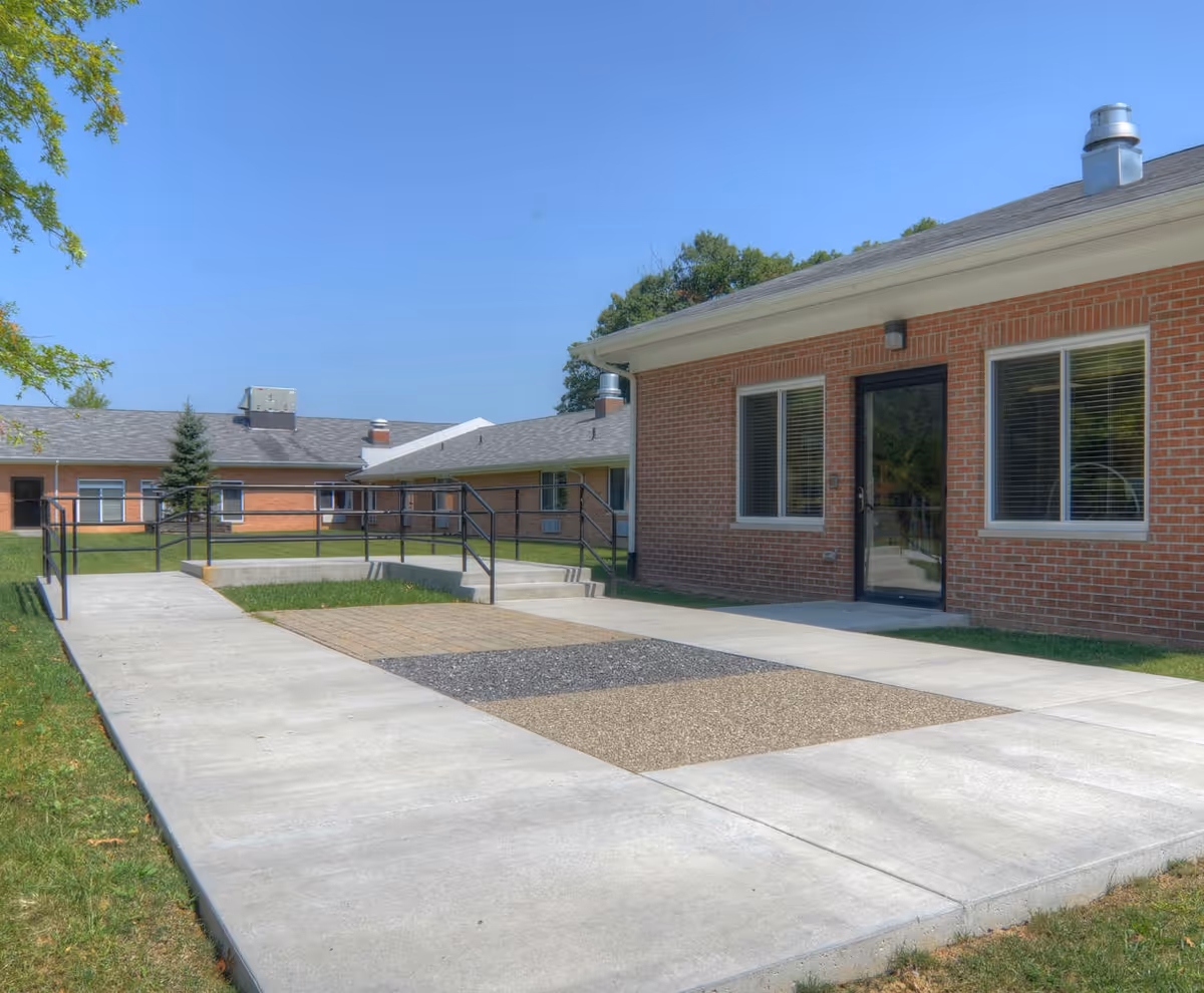 Brick single-story nursing facility exterior with a concrete walkway, wheelchair ramp, and windows under a clear blue sky.