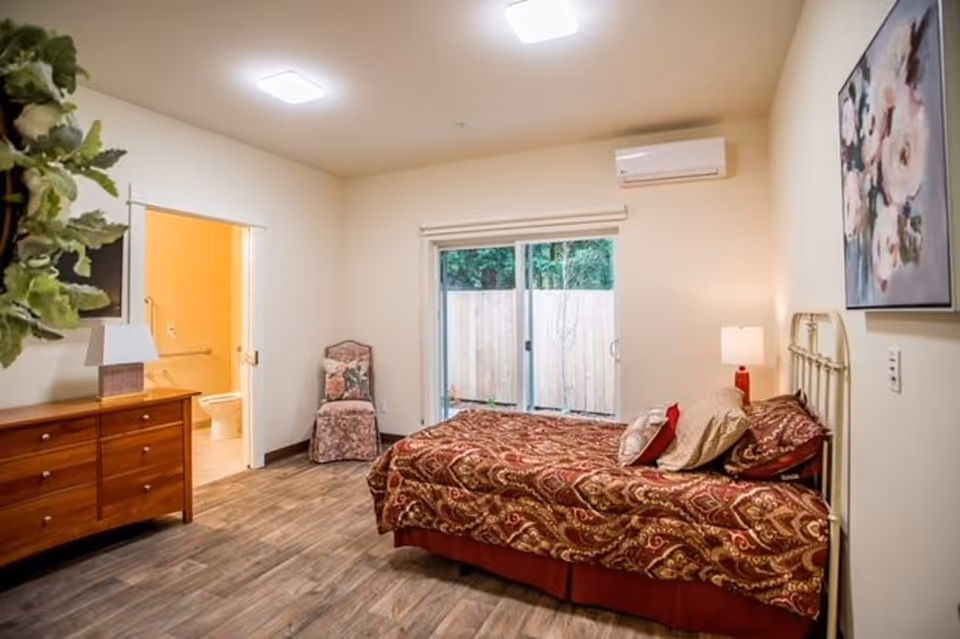 A furnished bedroom with a single bed, dresser, chair, sliding glass door and an open doorway to a bathroom.