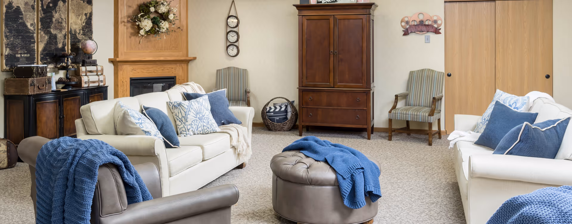 A cozy living room with two cream-colored sofas adorned with blue and patterned throw pillows and blankets. There is a round leather ottoman in the center with a blue knitted blanket draped over it. The room features a wooden cabinet, two striped armchairs, a fireplace with a floral wreath above it, and a wall clock with three faces. Decorative vintage suitcases and a globe are placed on a dark wooden sideboard, and a movie-themed wall decoration is visible near the wooden sliding doors.