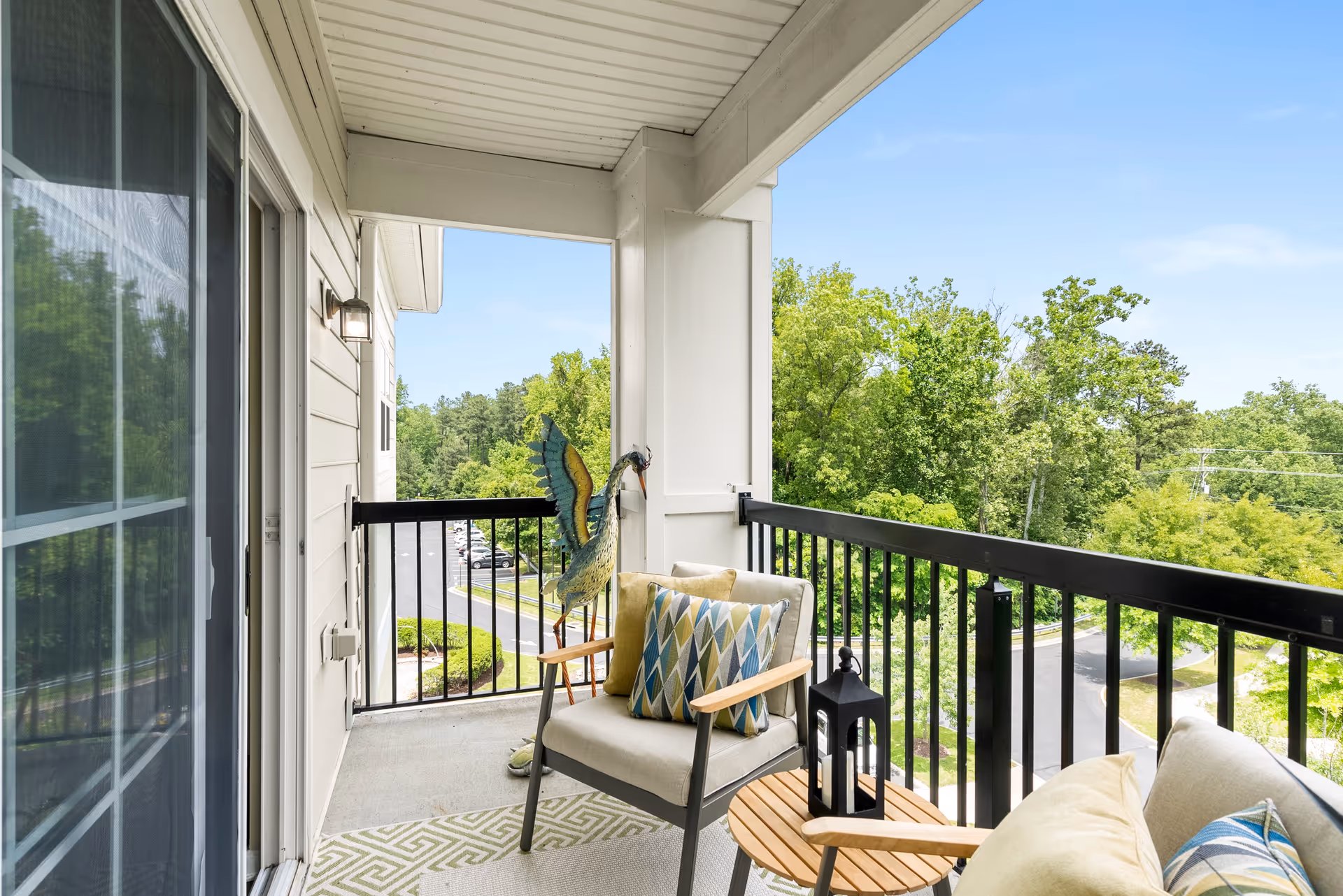 Covered balcony with cushioned chairs, a small round table, decorative pillows and a view of trees and a parking lot.