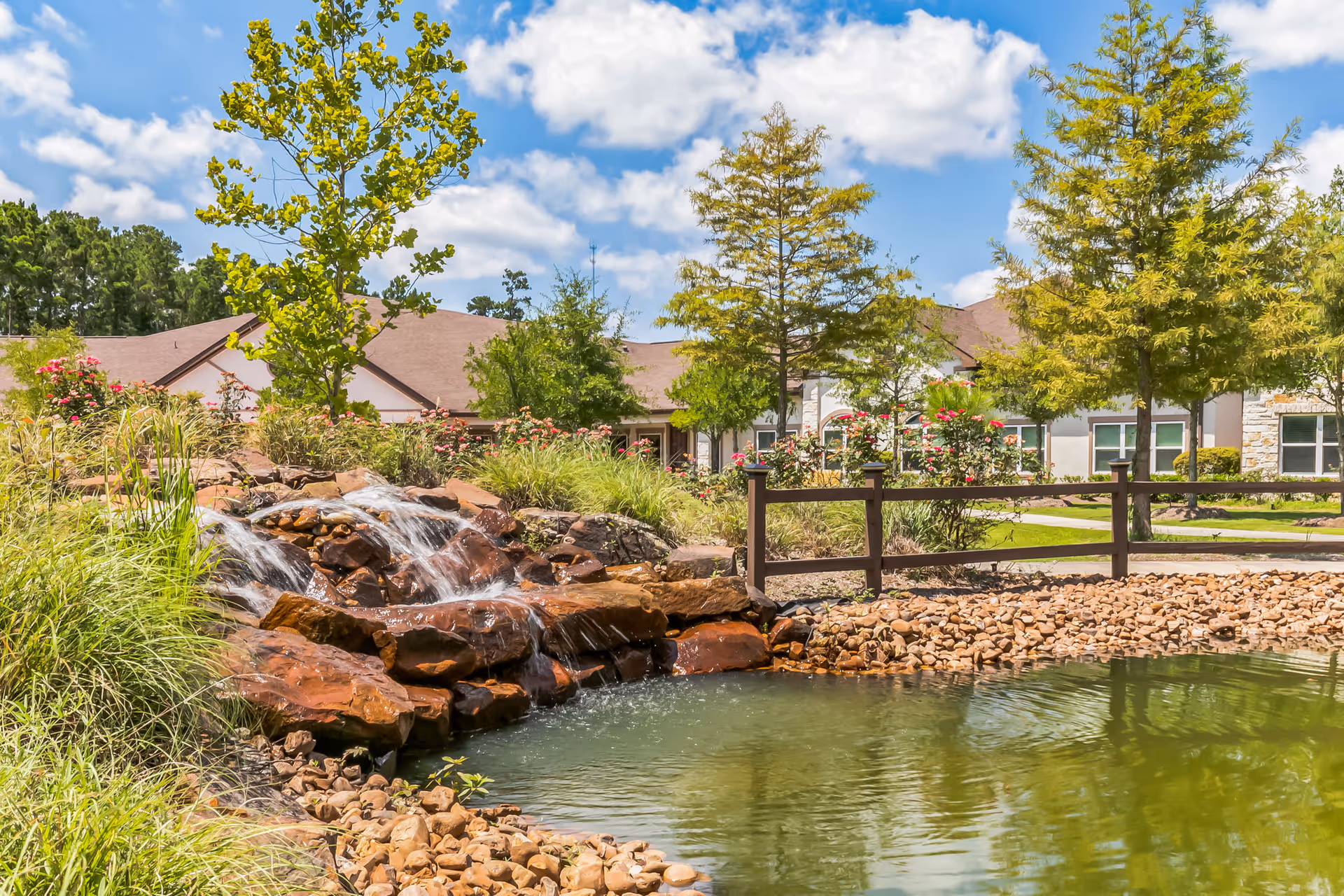 A landscaped outdoor area at Woodhaven Village featuring a small waterfall flowing over rocks into a pond, surrounded by green grass, trees, and flowering plants with a building in the background under a partly cloudy blue sky.