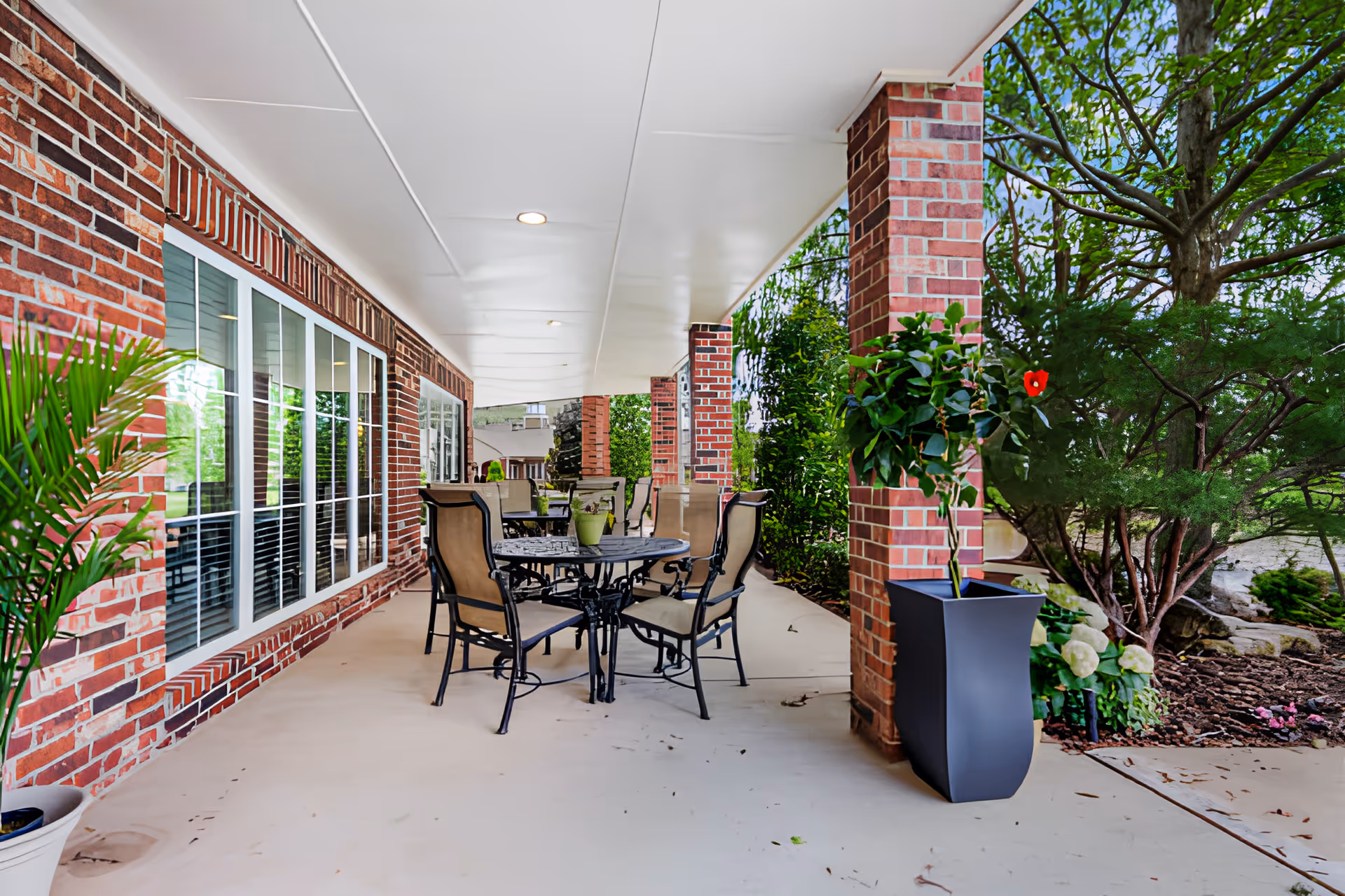 Covered outdoor patio with brick columns, a round metal table and several chairs, potted plants, and large windows along the building.