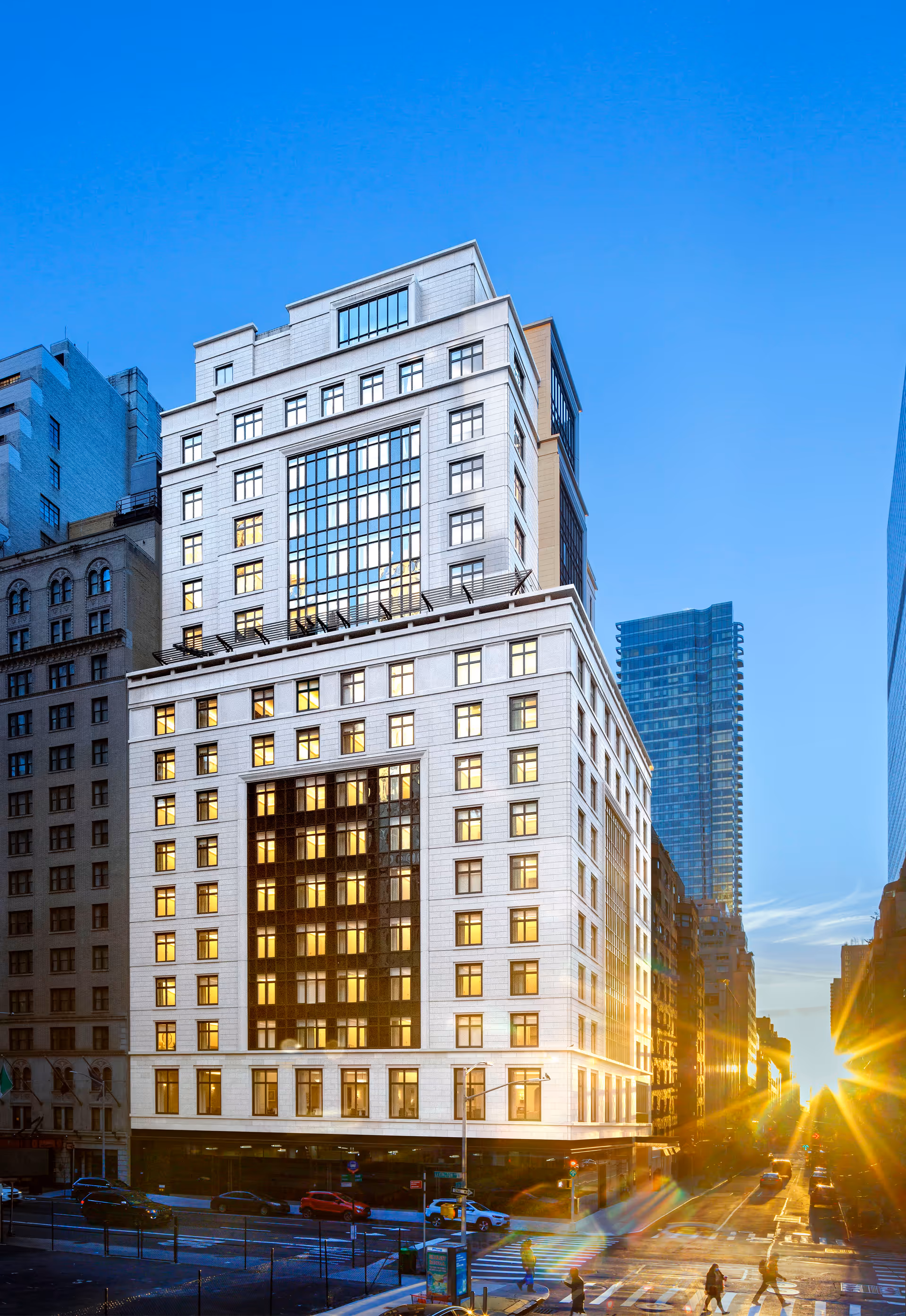 A tall, modern multi-story building with many windows reflecting sunlight, situated on a city street at sunset with people crossing the street and cars parked along the road.