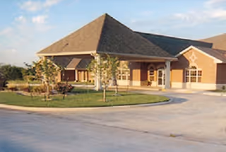 Exterior view of a single-story brick building with a sloped roof, surrounded by a small landscaped area with young trees and shrubs, and a paved driveway in front.