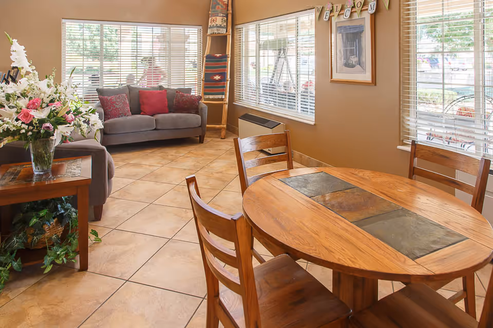 A cozy interior room with a round wooden table and four chairs in the foreground, a gray couch with red and patterned pillows near large windows with blinds, a side table with a vase of flowers, and a decorative ladder with blankets. A framed picture and a 'WELCOME' banner hang on the wall.