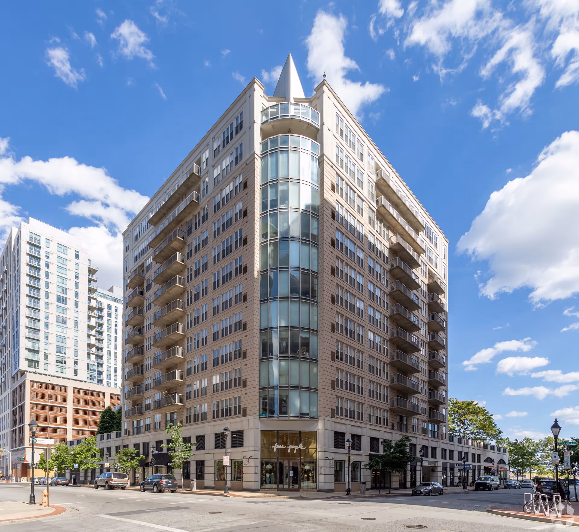 Exterior view of a multi-story modern building with large glass windows and balconies on a sunny day with blue sky and scattered clouds. The building is located at a street corner with parked cars and street lamps visible.