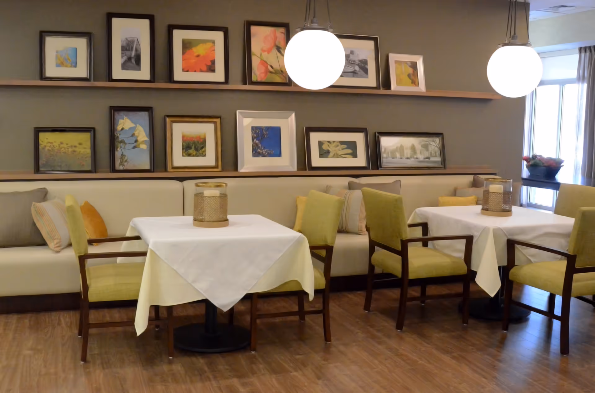 A cozy dining area with two tables covered in white tablecloths, surrounded by green cushioned chairs and a beige cushioned bench with colorful pillows. The wall behind features two wooden shelves displaying framed floral and nature artwork. Two round pendant lights hang from the ceiling, and a window with curtains allows natural light into the room.