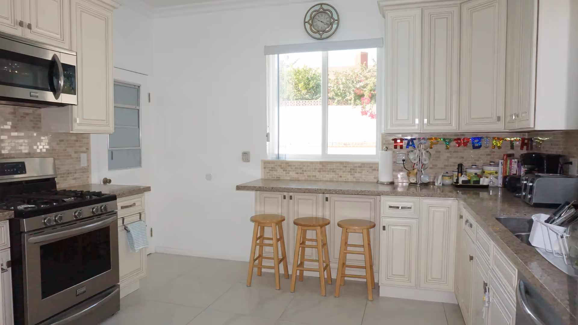 A bright kitchen with white cabinets, a stainless steel stove and microwave, a window above the counter, three wooden stools, and various kitchen appliances and items on the countertops. There is a colorful 'Happy Birthday' banner hanging on the wall under the cabinets.