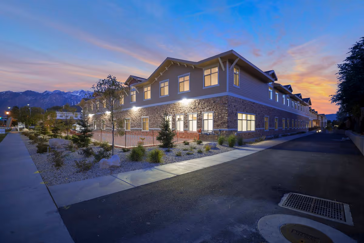 Exterior view of a two-story senior living facility building at dusk with lights on inside. The building has a stone and siding facade with multiple windows. There is a sidewalk and landscaped area with small trees and shrubs in front, and mountains are visible in the background under a colorful evening sky.