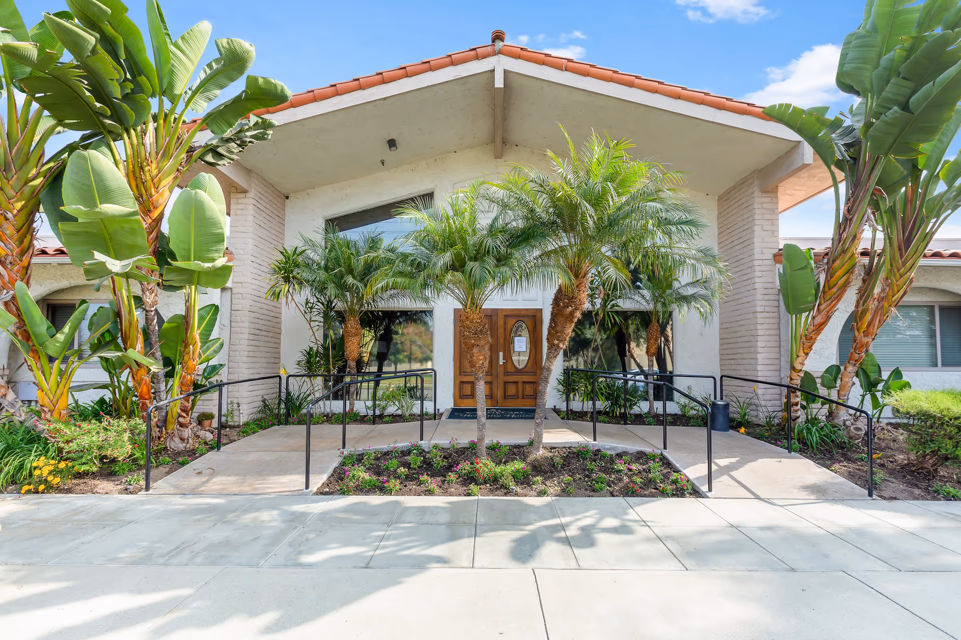 Front exterior view of Brittany House Residential Memory Care building with a peaked roof, large glass windows, wooden double doors, and landscaped garden featuring palm trees and tropical plants. There are concrete walkways with black handrails leading to the entrance.