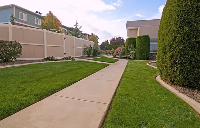 A paved walkway winding through a well-maintained grassy area with trimmed bushes and trees on either side, adjacent to residential buildings and a white fence under a partly cloudy sky.