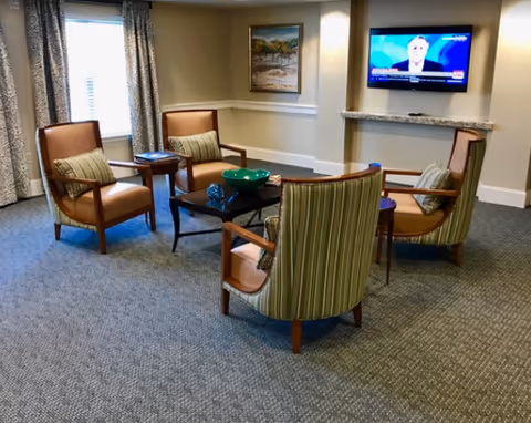 A cozy living room area with four armchairs arranged around a small black coffee table. The chairs have wooden frames with two featuring striped upholstery and two with solid brown cushions. There is a small side table between two chairs, a window with patterned curtains, a framed painting on the wall, and a flat-screen TV mounted on the wall showing a news broadcast.