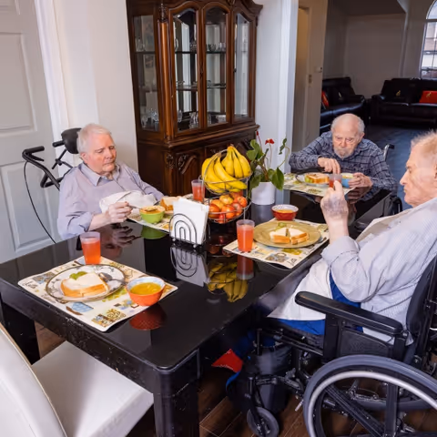 Three elderly men sitting around a black dining table in a well-lit room, eating a meal that includes sandwiches, soup, and juice. One man is in a wheelchair, and there is a centerpiece on the table with bananas, apples, and a small plant. A wooden cabinet with glass doors is visible in the background.