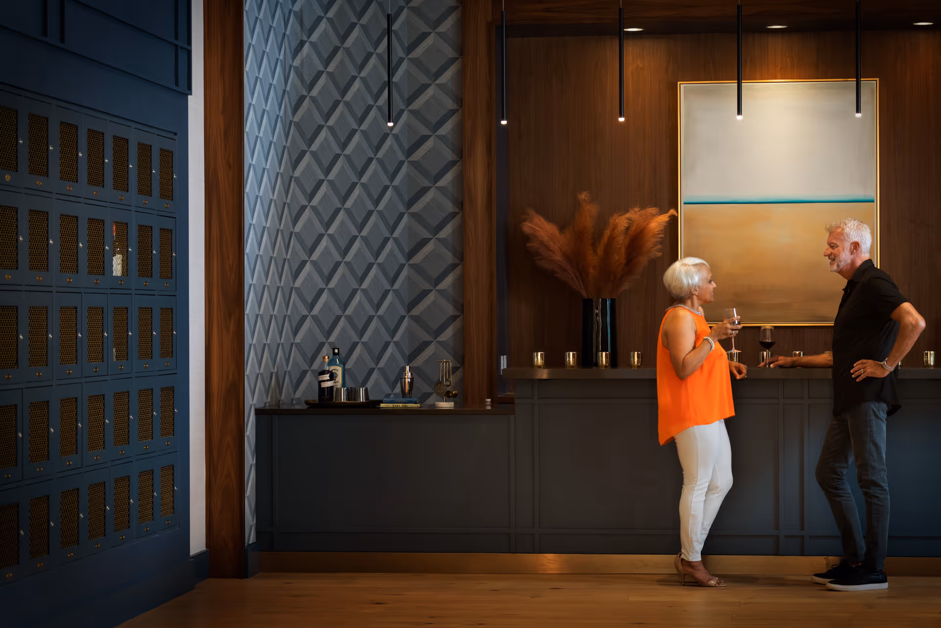 An older man and woman stand and chat while holding wine glasses in a stylish indoor bar area. The bar has a dark counter with decorative items including a vase with tall brown pampas grass and a large abstract painting on the wood-paneled wall behind them. To the left, there is a blue wall with multiple small lockers and a geometric patterned wall section. The floor is wooden, and several pendant lights hang from the ceiling.