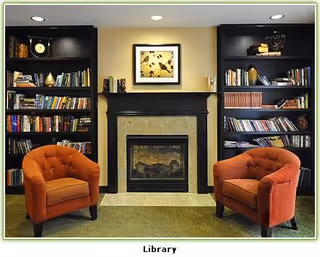 A cozy library area with two orange upholstered armchairs facing a fireplace. The fireplace is centered between two black bookshelves filled with books and decorative items. Above the fireplace hangs a framed picture.