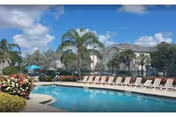 Outdoor swimming pool surrounded by lounge chairs and palm trees under a blue sky with some clouds. Residential buildings are visible in the background.